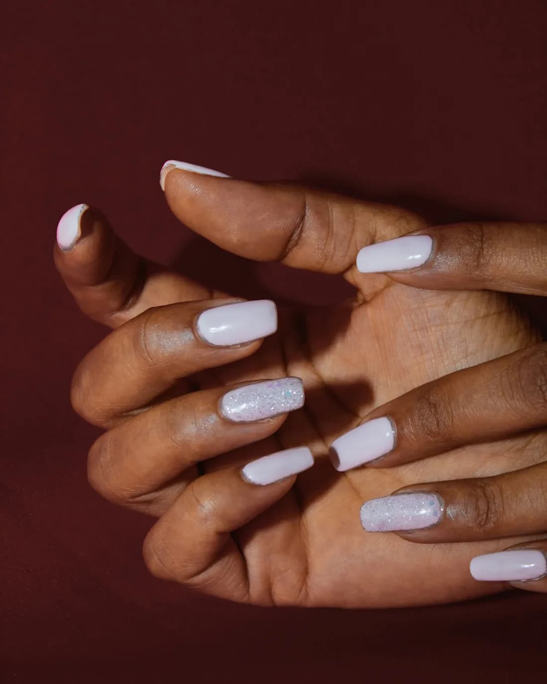 Two hands with pale lilac nails folded together on a burgundy backdrop