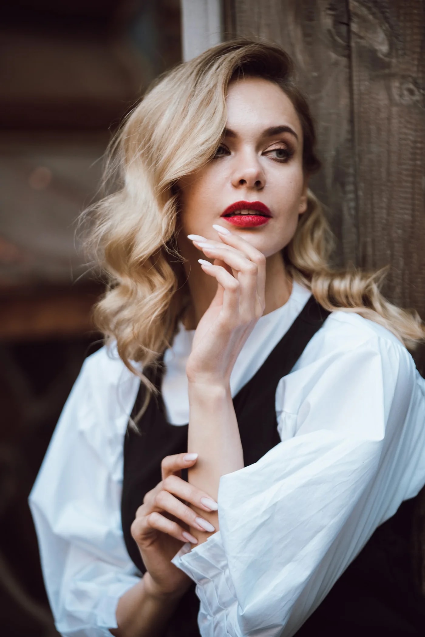 Woman with wavy blonde hair and red lips in a white blouse, hand raised to her chin