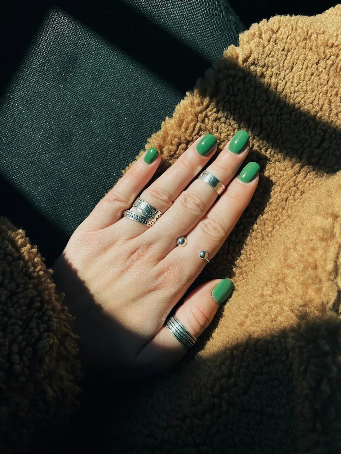A hand with green nails and silver rings resting on a shearling coat in sunlight